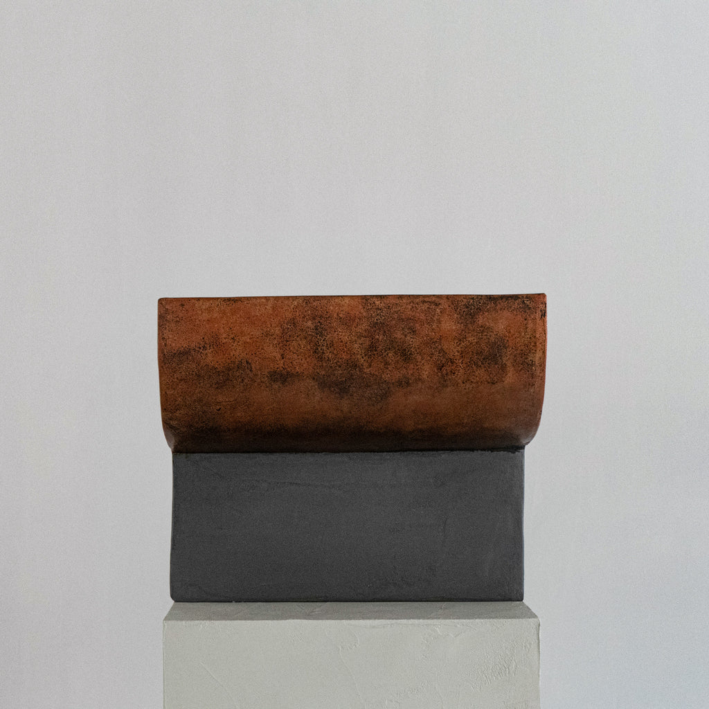 A sculptural stool finished in micro-cement and limestone. Sculpted into an abstract shape that features a curved, copper-toned metal half-shell resting over a matte, dark grey rectangular block. 