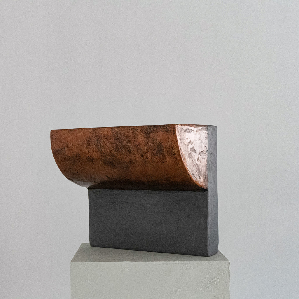 A sculptural stool finished in micro-cement and limestone. Sculpted into an abstract shape that features a curved, copper-toned metal half-shell resting over a matte, dark grey rectangular block. 