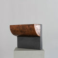 A sculptural stool finished in micro-cement and limestone. Sculpted into an abstract shape that features a curved, copper-toned metal half-shell resting over a matte, dark grey rectangular block. 