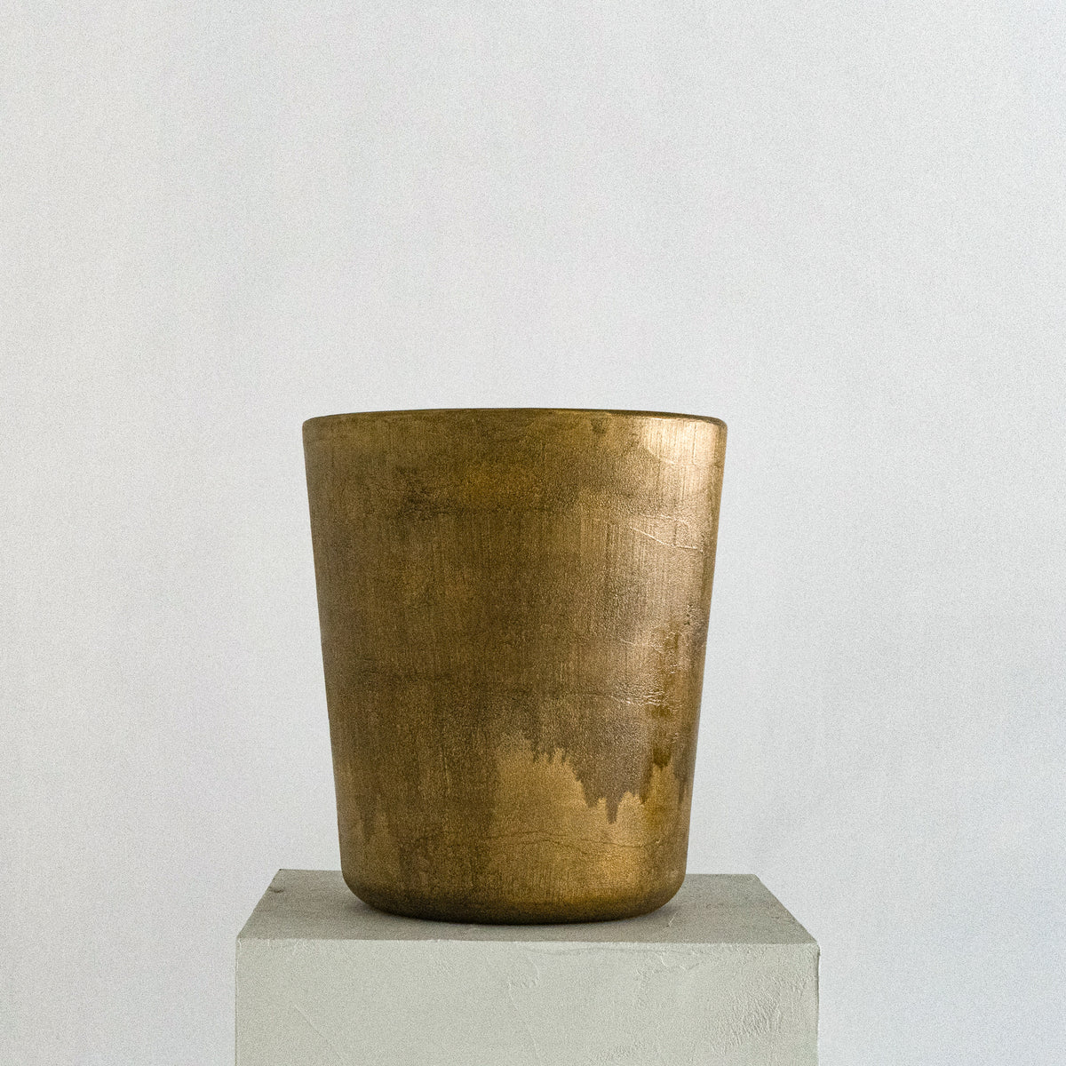 A cylindrical, metallic gold-finished Van Roosberg stool with a rough, brushed texture, displayed on a light gray concrete pedestal against a neutral off-white background.