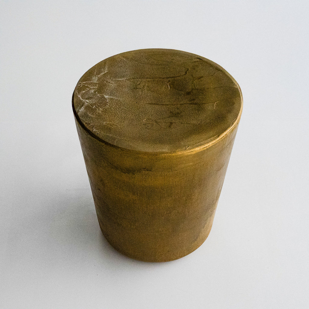 A cylindrical, metallic gold-finished Van Roosberg stool with a rough, brushed texture, displayed on a light gray concrete pedestal against a neutral off-white background.