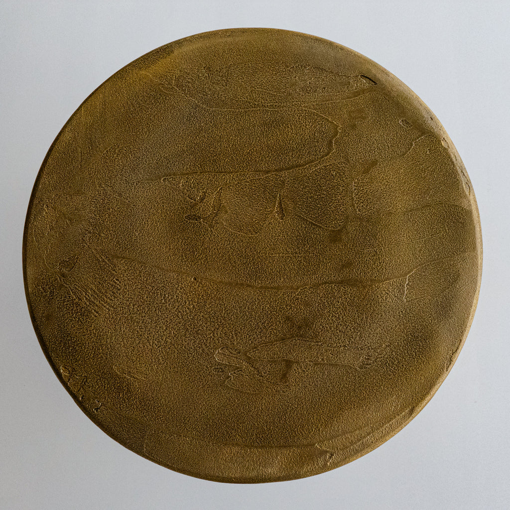 A cylindrical, metallic gold-finished Van Roosberg stool with a rough, brushed texture, displayed on a light gray concrete pedestal against a neutral off-white background.