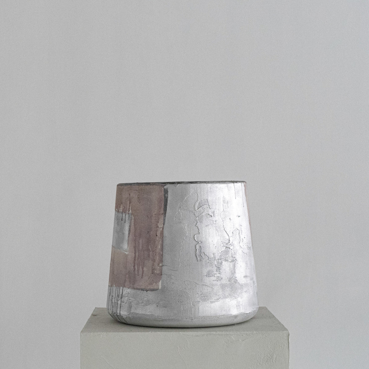 A handmade Van Roosberg sculptural stool with a tapered shape, featuring horizontal bands of textured dusty rose, metallic silver, and earthy brown glaze finishing. Displayed on a minimalist concrete pedestal against a neutral gray background.