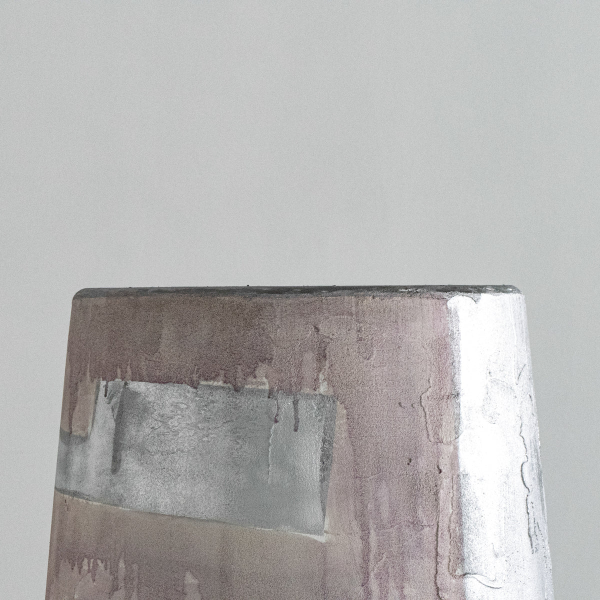 Close-up of the top edge of a textured rectangular stool with a distressed metallic finish in silver, taupe, and muted pink tones against a plain light gray background.