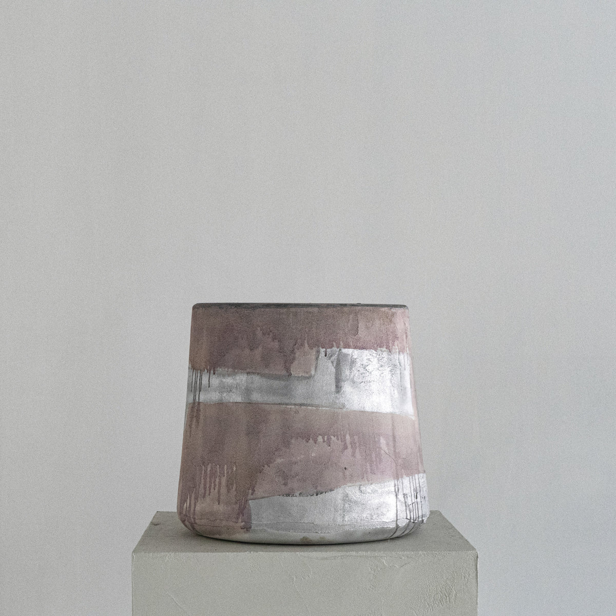 A handmade Van Roosberg sculptural stool with a tapered shape, featuring horizontal bands of textured dusty rose, metallic silver, and earthy brown glaze finishing. Displayed on a minimalist concrete pedestal against a neutral gray background.