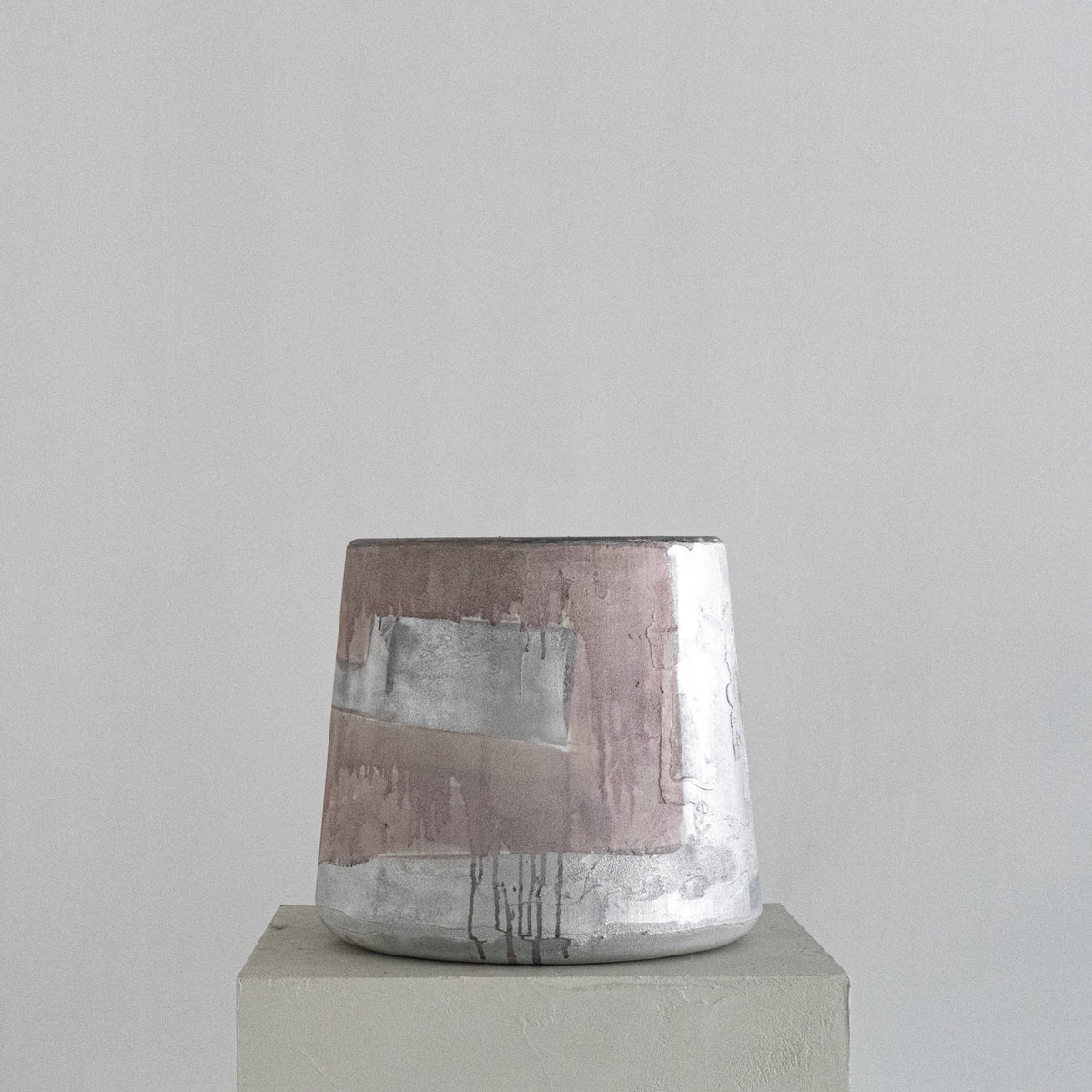 A handmade Van Roosberg sculptural stool with a tapered shape, featuring horizontal bands of textured dusty rose, metallic silver, and earthy brown glaze finishing. Displayed on a minimalist concrete pedestal against a neutral gray background.