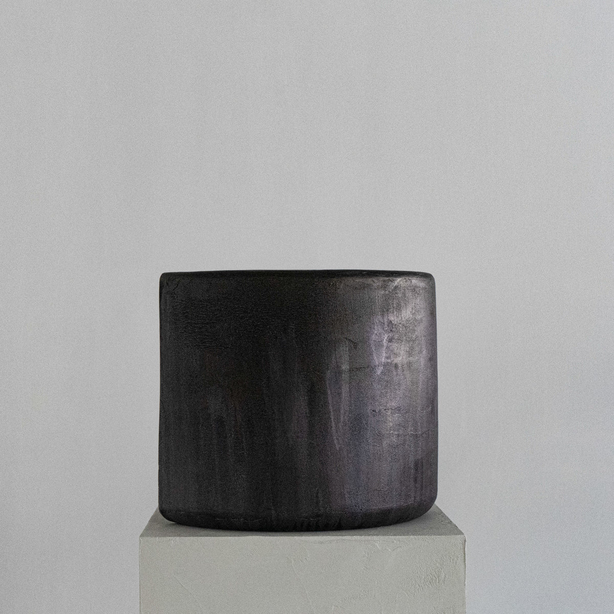A handmade sculptural cylindrical concrete stool with a textured, dual-toned finish in metallic silver and matte charcoal gray, displayed on a light gray pedestal against a neutral gray background.