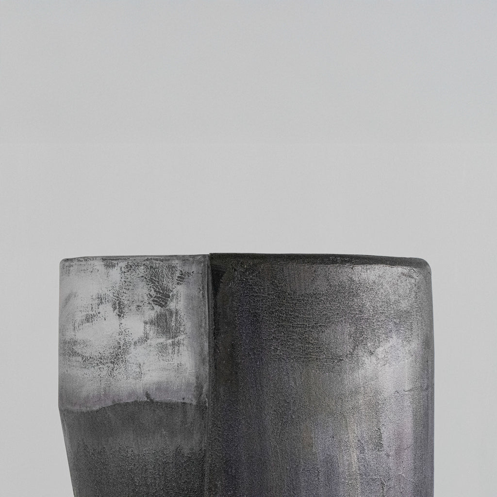 A handmade sculptural cylindrical concrete stool with a textured, dual-toned finish in metallic silver and matte charcoal gray, displayed on a light gray pedestal against a neutral gray background.