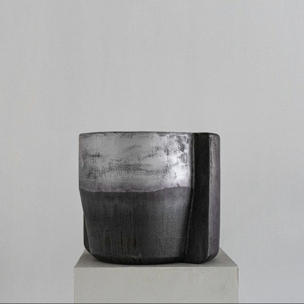 A handmade sculptural cylindrical concrete stool with a textured, dual-toned finish in metallic silver and matte charcoal gray, displayed on a light gray pedestal against a neutral gray background.