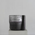 A handmade sculptural cylindrical concrete stool with a textured, dual-toned finish in metallic silver and matte charcoal gray, displayed on a light gray pedestal against a neutral gray background.