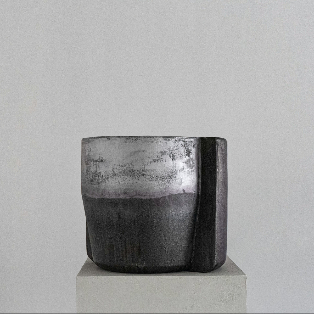 A handmade sculptural cylindrical concrete stool with a textured, dual-toned finish in metallic silver and matte charcoal gray, displayed on a light gray pedestal against a neutral gray background.