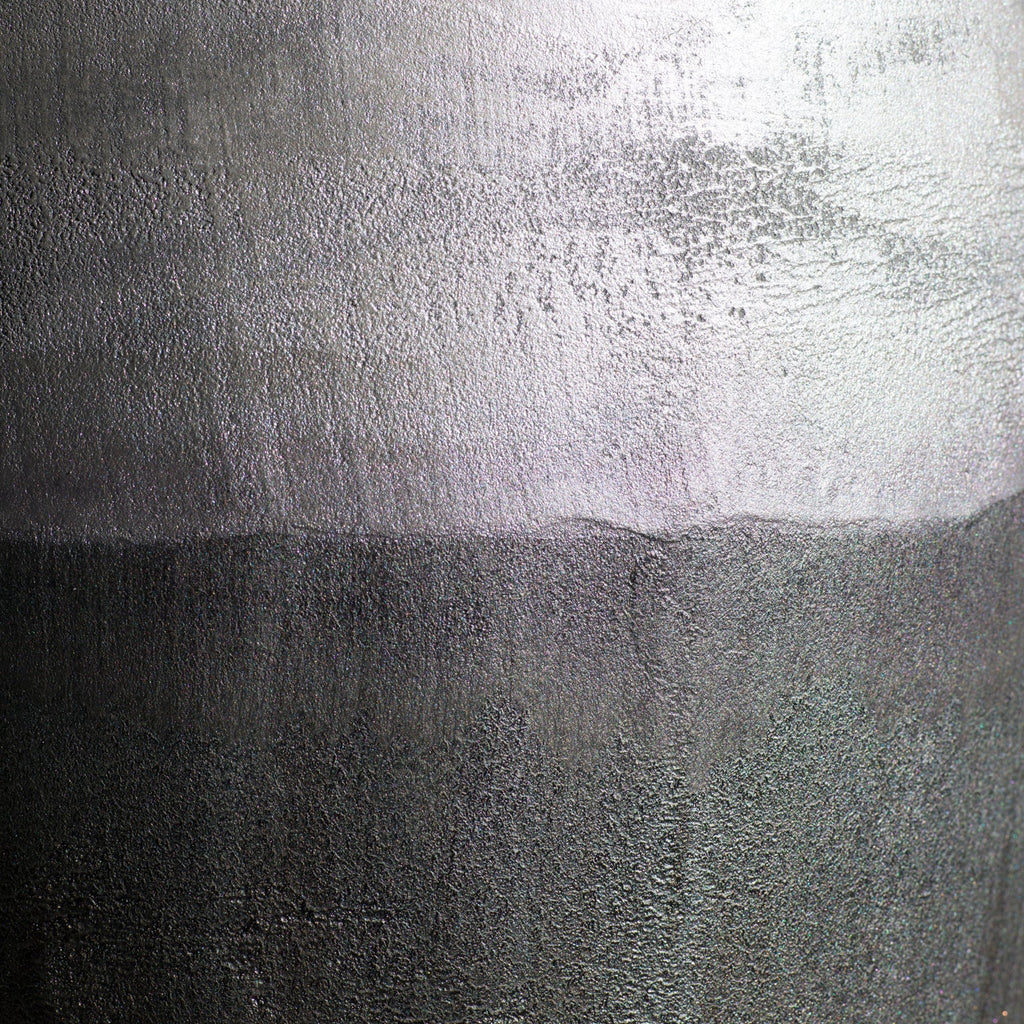 A close-up of a Van Roosberg metallic-grey stool with a textured metallic surface with a horizontal color transition, showing a bright silver upper section and a darker gunmetal gray lower section.