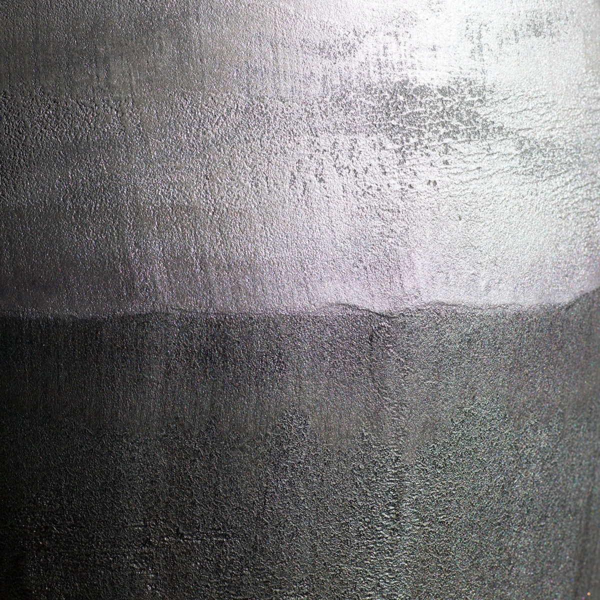 A close-up of a Van Roosberg metallic-grey stool with a textured metallic surface with a horizontal color transition, showing a bright silver upper section and a darker gunmetal gray lower section.