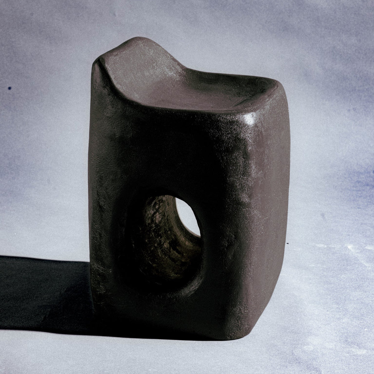 A sculptural stool finished in micro-cement (limestone) with natural pigments in a deep charcoal-brown tone. Its rounded rectangular form rises into an asymmetric seat with a subtle backrest, while a smooth oval cutout pierces the front, opening a window through the piece. A hand-finished texture gives it a raw, sculptural presence.