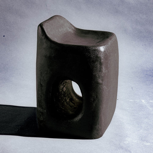A sculptural stool finished in micro-cement (limestone) with natural pigments in a deep charcoal-brown tone. Its rounded rectangular form rises into an asymmetric seat with a subtle backrest, while a smooth oval cutout pierces the front, opening a window through the piece. A hand-finished texture gives it a raw, sculptural presence.