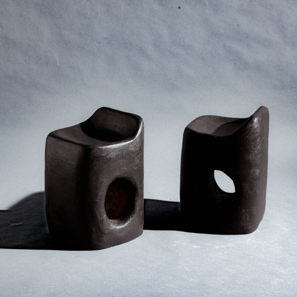 Two sculptural stool finished in micro-cement (limestone) with natural pigments in a deep charcoal-brown tone. Its rounded rectangular form rises into an asymmetric seat with a subtle backrest, while a smooth oval cutout pierces the front, opening a window through the piece. A hand-finished texture gives it a raw, sculptural presence.