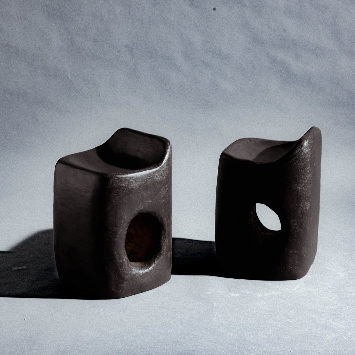Two sculptural stool finished in micro-cement (limestone) with natural pigments in a deep charcoal-brown tone. Its rounded rectangular form rises into an asymmetric seat with a subtle backrest, while a smooth oval cutout pierces the front, opening a window through the piece. A hand-finished texture gives it a raw, sculptural presence.
