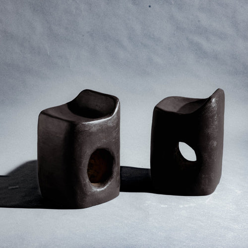 Two sculptural stool finished in micro-cement (limestone) with natural pigments in a deep charcoal-brown tone. Its rounded rectangular form rises into an asymmetric seat with a subtle backrest, while a smooth oval cutout pierces the front, opening a window through the piece. A hand-finished texture gives it a raw, sculptural presence.