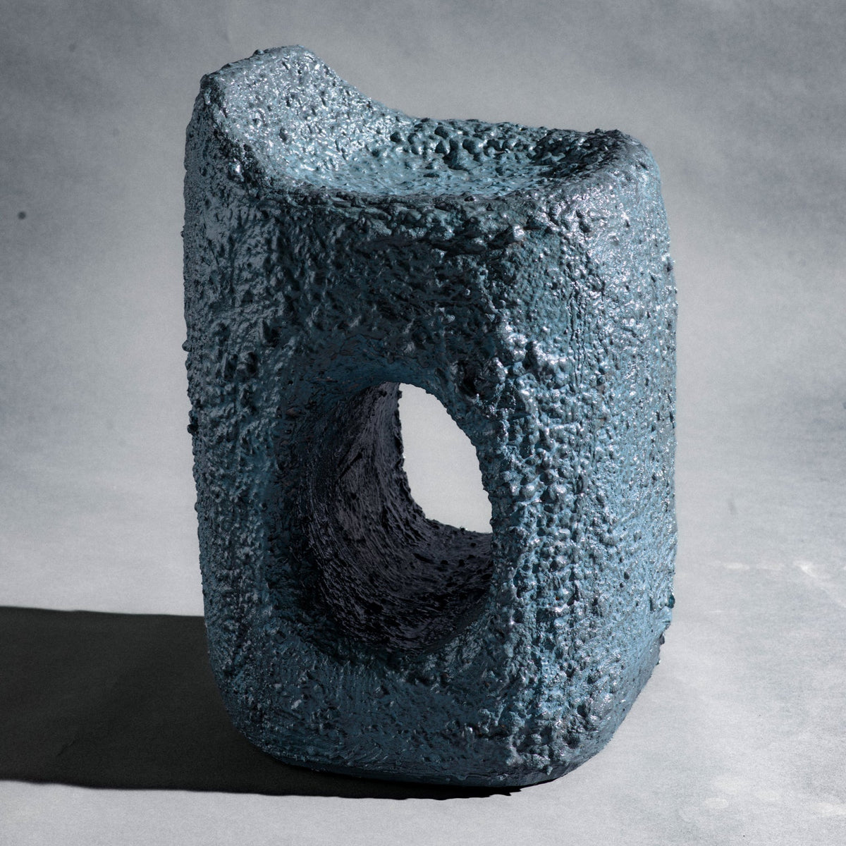 A sculptural stool finished in micro-cement (limestone) with natural pigments in a dark teal-blue tone and a rough, stone-like texture. Its rounded rectangular form rises into an asymmetric seat with a subtle backrest, while a smooth central hole pierces the front, opening a window through the piece. The hand-finished surface gives it a raw, abstract, sculptural presence.