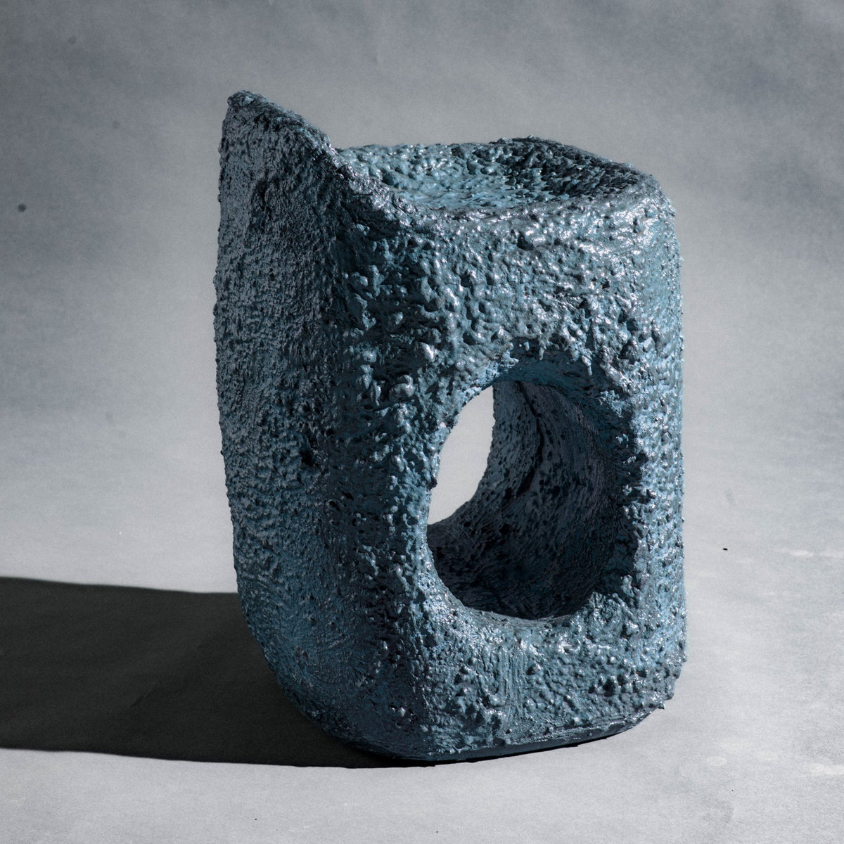 A sculptural stool finished in micro-cement (limestone) with natural pigments in a dark teal-blue tone and a rough, stone-like texture. Its rounded rectangular form rises into an asymmetric seat with a subtle backrest, while a smooth central hole pierces the front, opening a window through the piece. The hand-finished surface gives it a raw, abstract, sculptural presence.