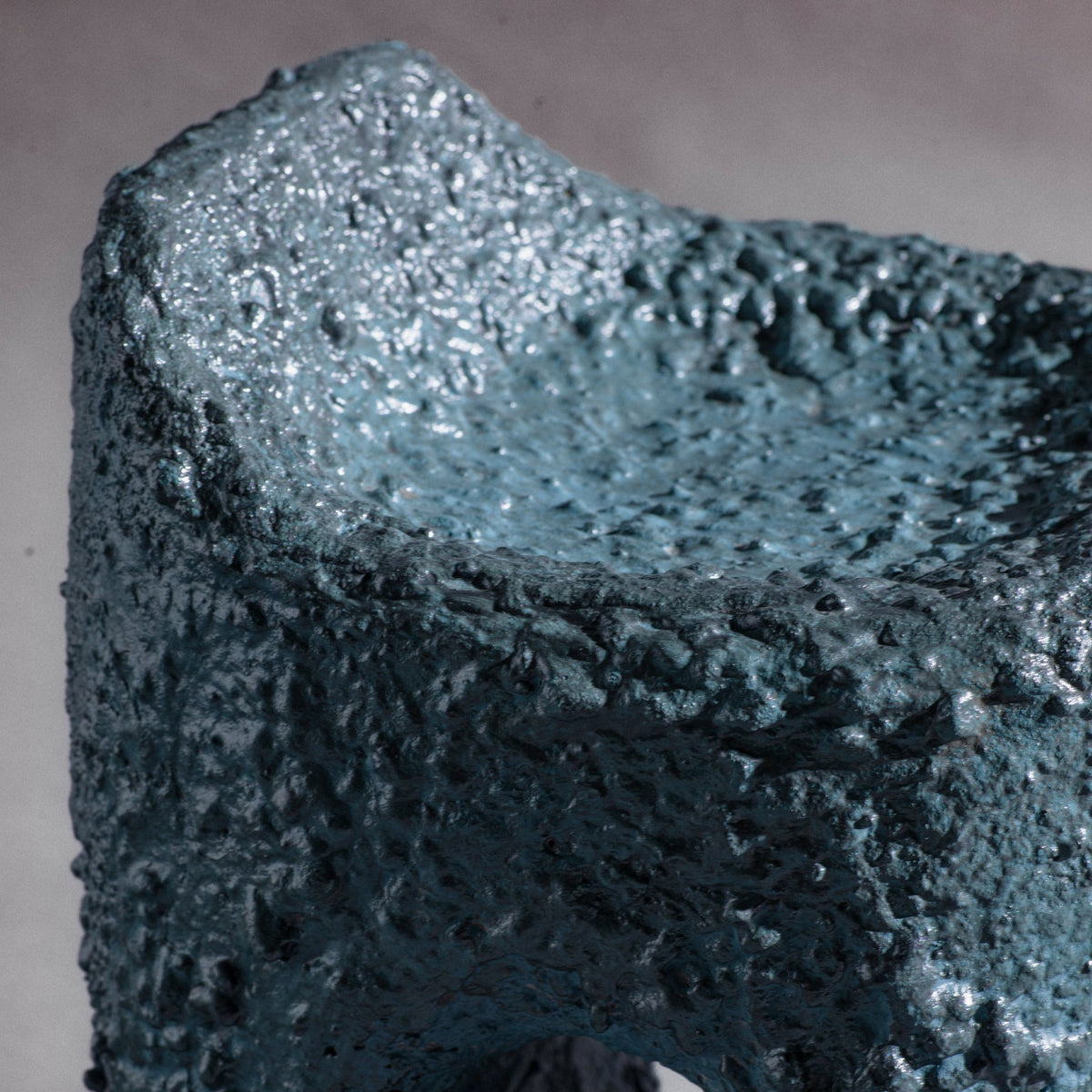 A sculptural stool finished in micro-cement (limestone) with natural pigments in a dark teal-blue tone and a rough, stone-like texture. Its rounded rectangular form rises into an asymmetric seat with a subtle backrest, while a smooth central hole pierces the front, opening a window through the piece. The hand-finished surface gives it a raw, abstract, sculptural presence.