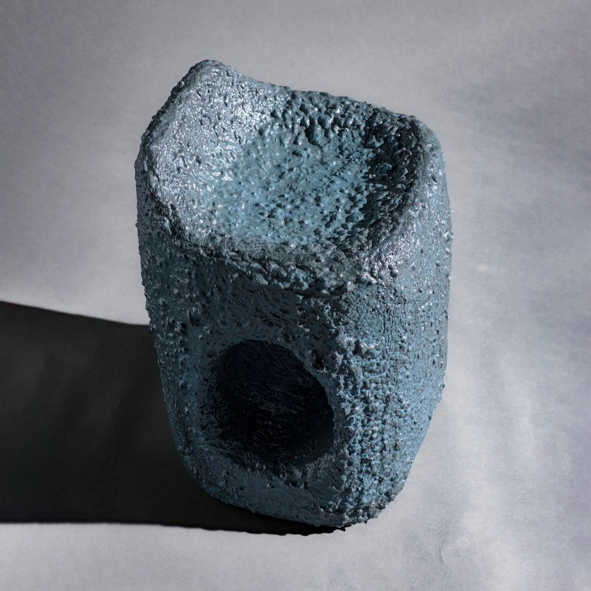 A sculptural stool finished in micro-cement (limestone) with natural pigments in a dark teal-blue tone and a rough, stone-like texture. Its rounded rectangular form rises into an asymmetric seat with a subtle backrest, while a smooth central hole pierces the front, opening a window through the piece. The hand-finished surface gives it a raw, abstract, sculptural presence.