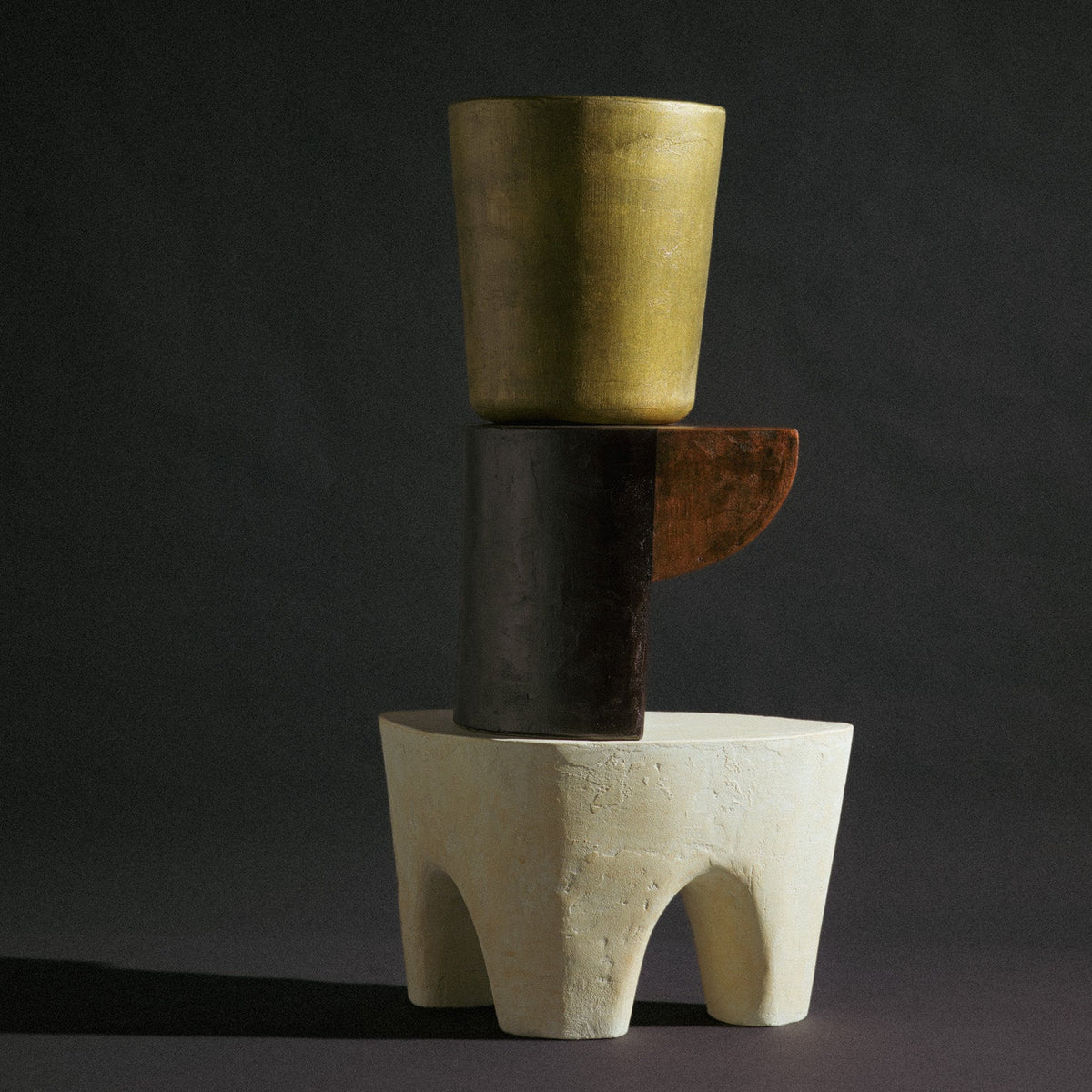 An abstract sculptural arrangement of stacked geometric forms on a dark studio background: a matte gold cylindrical vessel on top, a dark charcoal cylinder with a rust-brown side tab, and a textured off-white pedestal base.