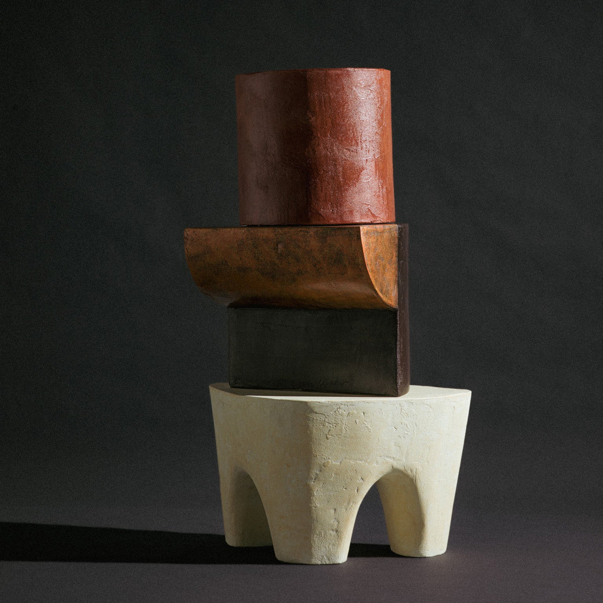A trio of abstract sculptural stools by Van Roosberg, stacked to form one decorative statement piece. At the base sits a beige, three-legged stool shaped like an elephant's rounded body, grounding the composition. Stacked on top is a copper stool in a reversed L-shape, its angular silhouette contrasting the organic base below. Crowning the stack is a cylindrical stool in a blood-orange hue that fades into dark grey toward its base. Each stool holds its own distinct color and form, yet together they read as 