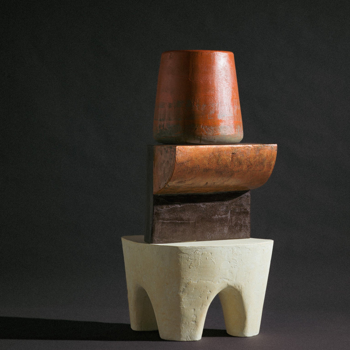 A trio of abstract sculptural stools by Van Roosberg, stacked to form one decorative statement piece. At the base sits a beige, three-legged stool shaped like an elephant's rounded body, grounding the composition. Stacked on top is a copper stool in a reversed L-shape, its angular silhouette contrasting the organic base below. Crowning the stack is a cylindrical stool in a blood-orange hue that fades into dark grey toward its base. Each stool holds its own distinct color and form, yet together they read as 