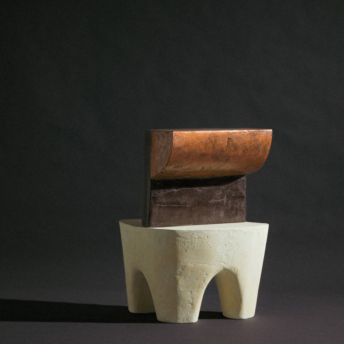 An abstract shape sculptural copper stool by Van Roosberg stacked on top of beige 3 legged stool that has elephant body shape. Two stool stacked on top of each other against dark studio background.