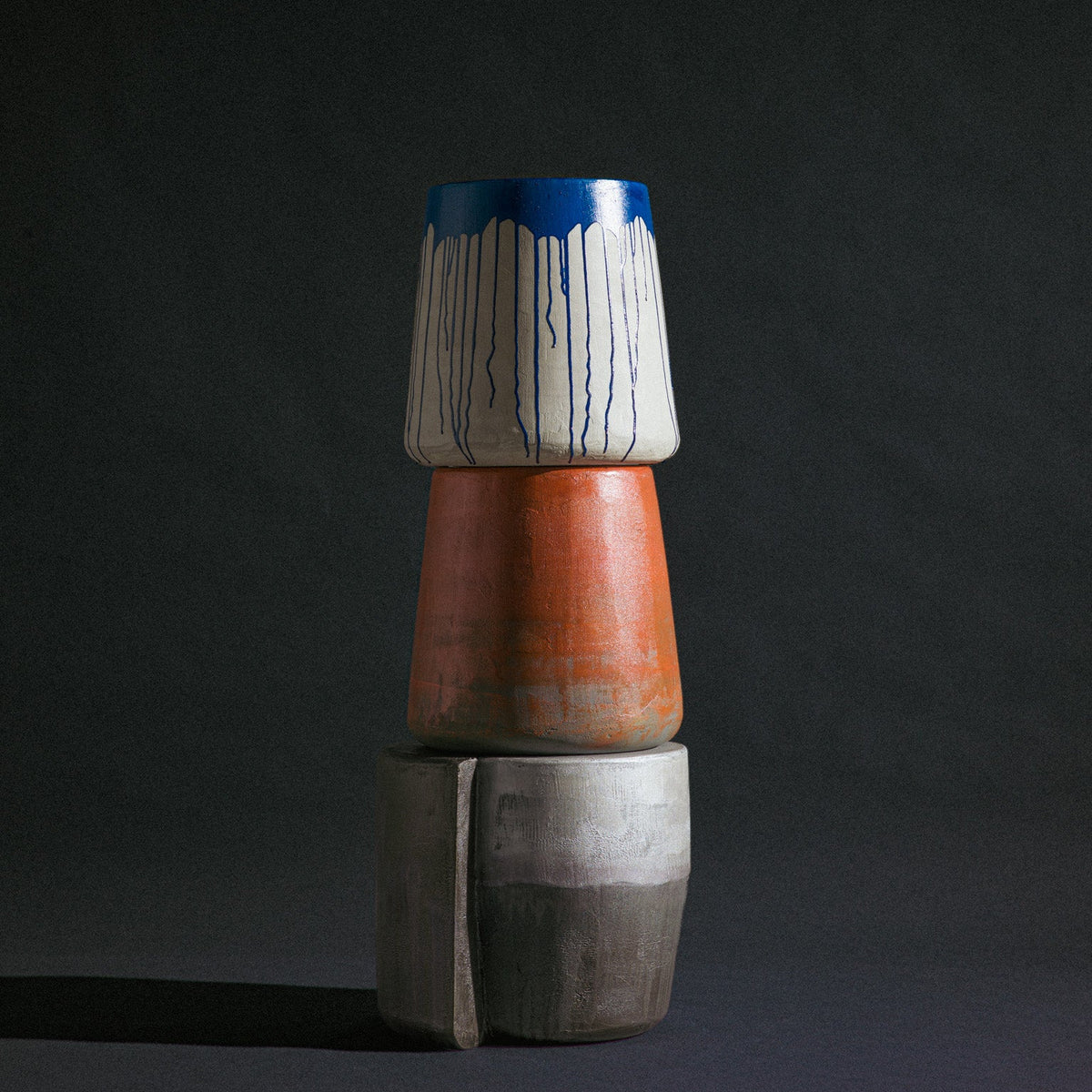 A trio of abstract sculptural stools by Van Roosberg, stacked to form one decorative statement piece. At the base sits a silver grey stool grounding the composition. Stacked on top is a blood orange cylindrical stool, its bright color stand out. Crowning the stack is a cylindrical stool in light grey finishing with bright blue painting dripping down from the top. Each stool holds its own distinct color and form.